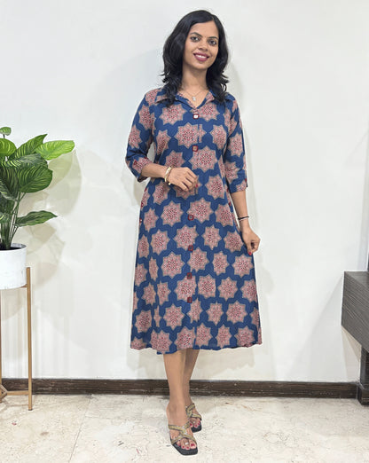 Blue women's Ajrakh vegetable dyed plant based cotton Long kurti with a blue base & abstract red Star print, standing indoors with a white wall and plant in the background