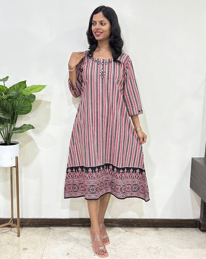 Red Woman's Ajrakh Pure cotton long-kurti with a strip print, ajrakh print at bottom, Standing in front of white wall and plant in the background.