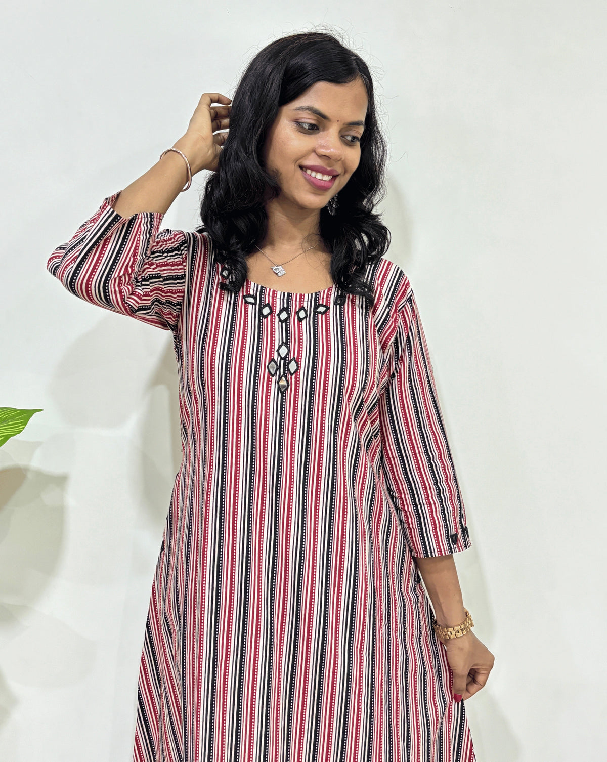 Close-up of Red Woman's Ajrakh Pure cotton long-kurti with a Multi-strip print with black, Red & Beige, ajrakh print at bottom, Hand-mirror work on front, Sleeves cuffs & Round neck, Standing in front of white wall and plant in the background. 