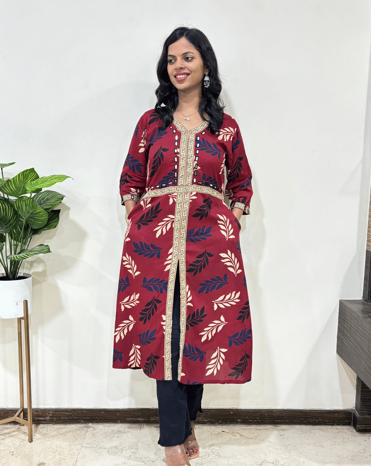 Front view of Red Woman's Ajrakh Vegetable dyed Planet based cotton Long-kurti with a leaf print along with Beige Ajrakh print on border from V-shaped neck on front & Sleeves cuffs with Middle Slit, 2-side pocket,  standing indoors with a plant and white wall in the background.