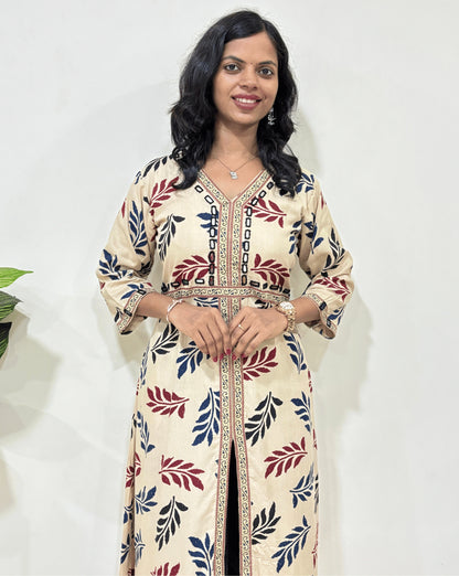Close-up of Beige Woman's Ajrakh Vegetable dyed Planet based cotton Long-kurti with a leaf print along with Ajrakh print & Hand-mirror work on border from V-shaped neck on front & Sleeves cuffs with Middle Slit, standing indoors with a plant and white wall in the background.