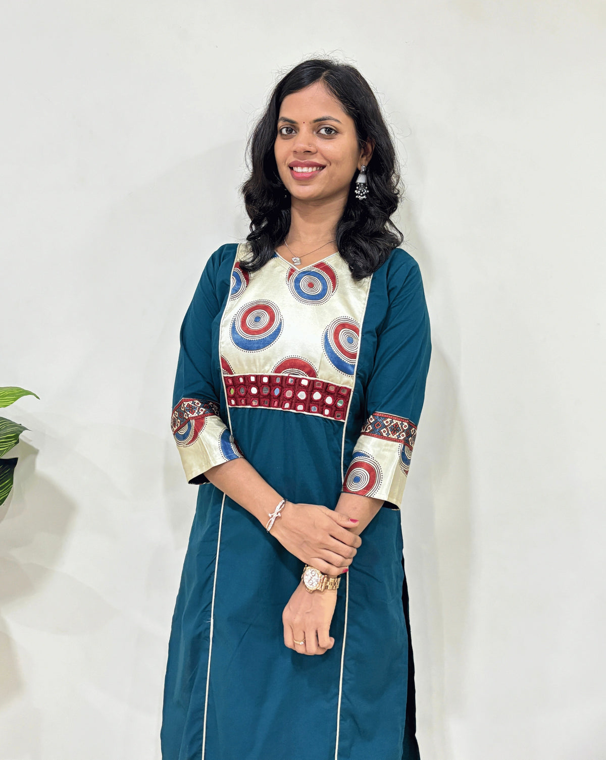 Close-up of Teal-Green Woman's Ajrakh Vegetable Dyed Plant Based Rayon Cotton Long-Kurti Featured with Three-Quarter Sleeves with White Half Mashru silk Geometric yoke Pattern on front &  Sleeves cuffs, Hand-mirror work on front with white strip panel, Paired with Black jeans, Standing in Front of White Wall, Green plant Pot on left side.