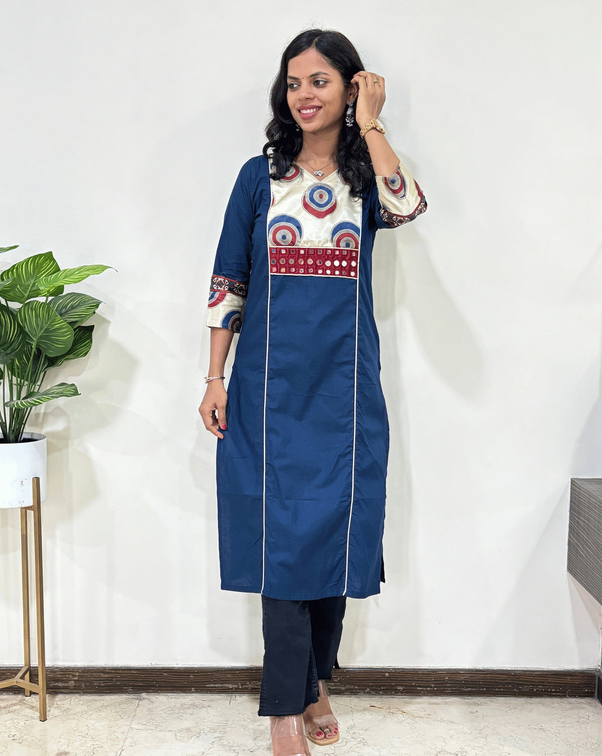 Deep Blue Woman's Ajrakh Vegetable Dyed Plant Based Rayon Cotton Long-Kurti with White Half Mashru silk Geometric yoke Pattern on front Sleeves cuffs, Paired with Black jeans, Standing in Front of White Wall, Green plant Pot on left side.
