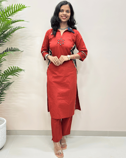 Front view of Ajrakh woman's vegetable dyed cotton co-ord set with black and red embroidered details, v-shaped collar neck, hand-mirror work on chest , sleeves, standing against a beige wall with a green plant on the left.