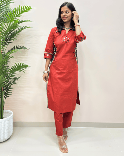 Red women's ajrakh kurta set with matching pants, featuring mirror embroidery & ajrakh pattern with semi-moon print,  standing in a room with a potted plant on the left.