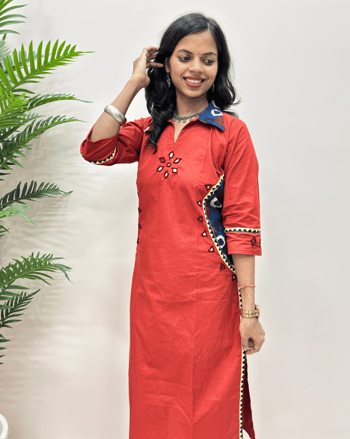 Side-veiw of red cotton Co-ord set  with blue, black, and beige embroidered details, standing against a light beige wall with a green plant on the left.