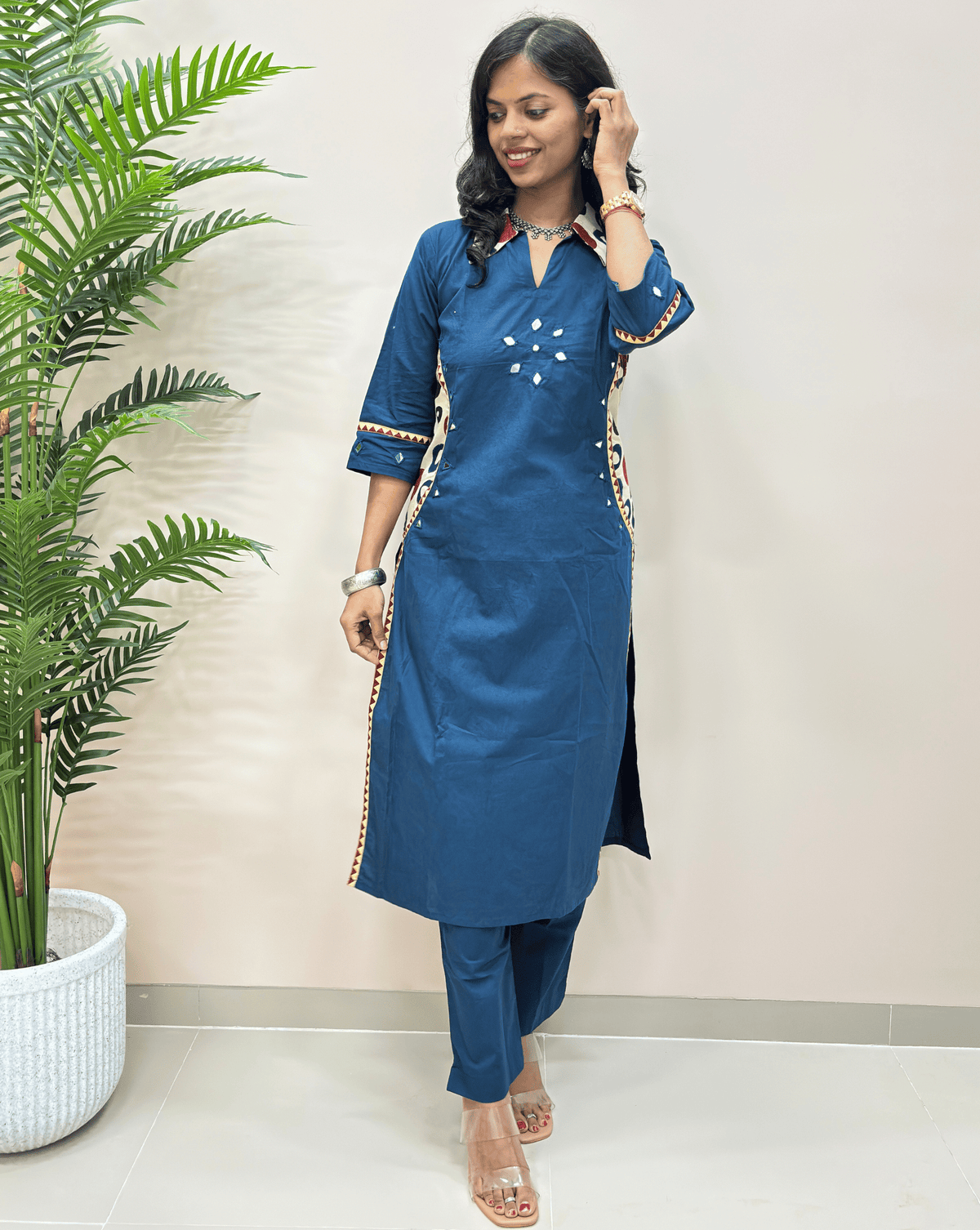 Blue women's plant based cotton long-kurti with pant with beige and white decorative geometric borders and embroidery, V-shaped collar-neck standing in front of a beige wall next to a potted green plant.