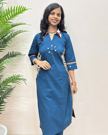 Close-up of blue ajrakh vegetable dyed cotton Co-ord set with decorative white and beige embroidery with geometric prints, standing against a plain light beige wall with a green plant on the left.