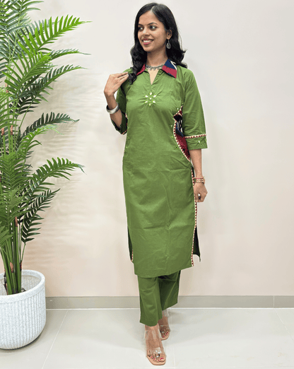  Green women's ajrakh long cotton co-ord set, featuring a red and navy collar, mirror embroidery on the chest, and beige trim on the sleeve cuffs, standing in front of a beige wall next to a potted plant.