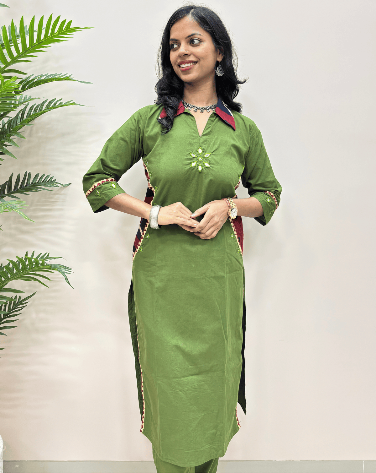 Close-up of green ajrakh vegetable dyed cotton Co-ord set with red and beige trim details, mirror work standing against a plain light background with a green plant on the left.