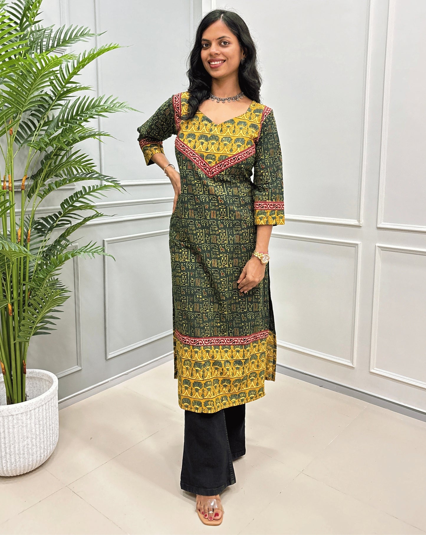 Dark green women's ajrakh hand-block pure cotton long-kurti featured with yellow base & green abstract ajrakh print, paired with black flared jeans. standing in front of white wall.