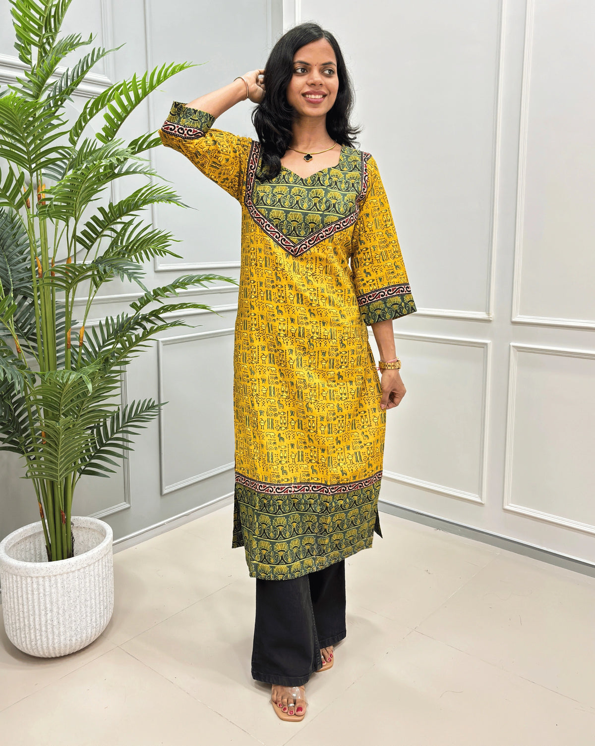  yellow and green women's hand-block ajrakh pure cotton printed long- kurti,  paired with black flared jeans , standing in a bright indoor setting with a potted plant and paneled wall.