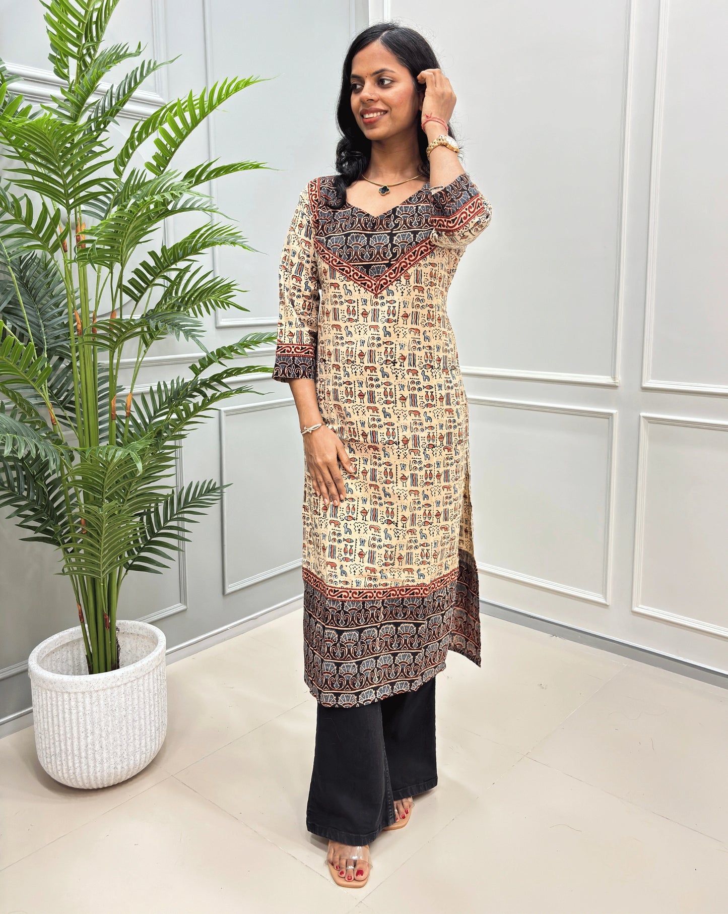 Beige and Black women's hand-block ajrakh pure cotton printed long- kurti,  paired with black flared jeans , standing in a bright indoor setting with a potted plant and paneled wall.