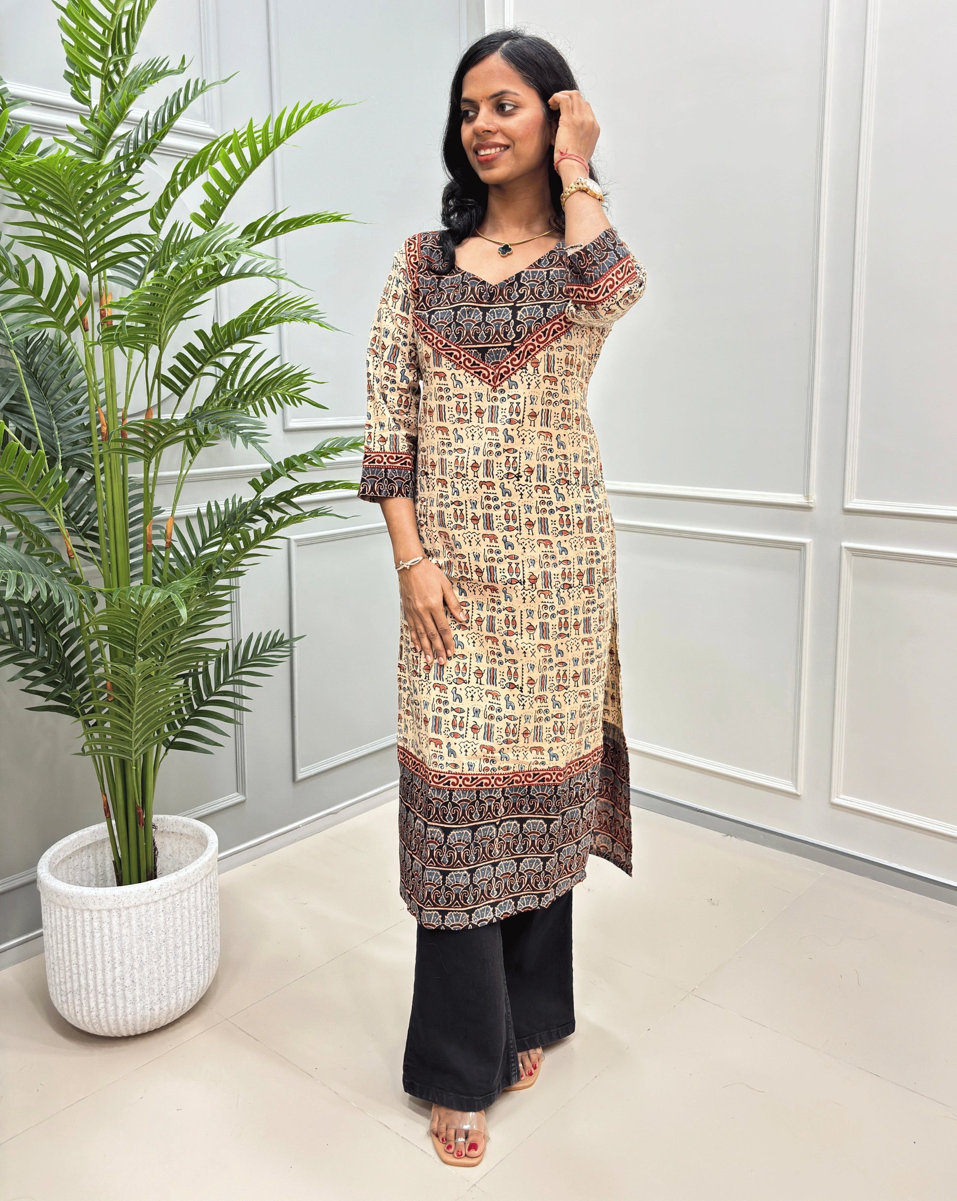 Beige and Black women's hand-block ajrakh pure cotton printed long- kurti,  paired with black flared jeans , standing in a bright indoor setting with a potted plant and paneled wall.