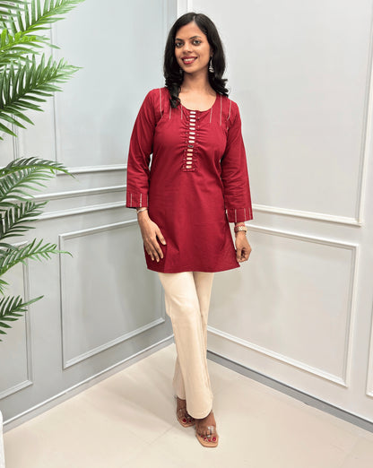 Deep Red women's Ajrakh Vegetable Dyed plant based Cotton Short-kurti with white piping and Hand-mirror work look a like button details, paired with beige Flared pants, standing in a room with a light gray paneled wall and a green plant on the left.