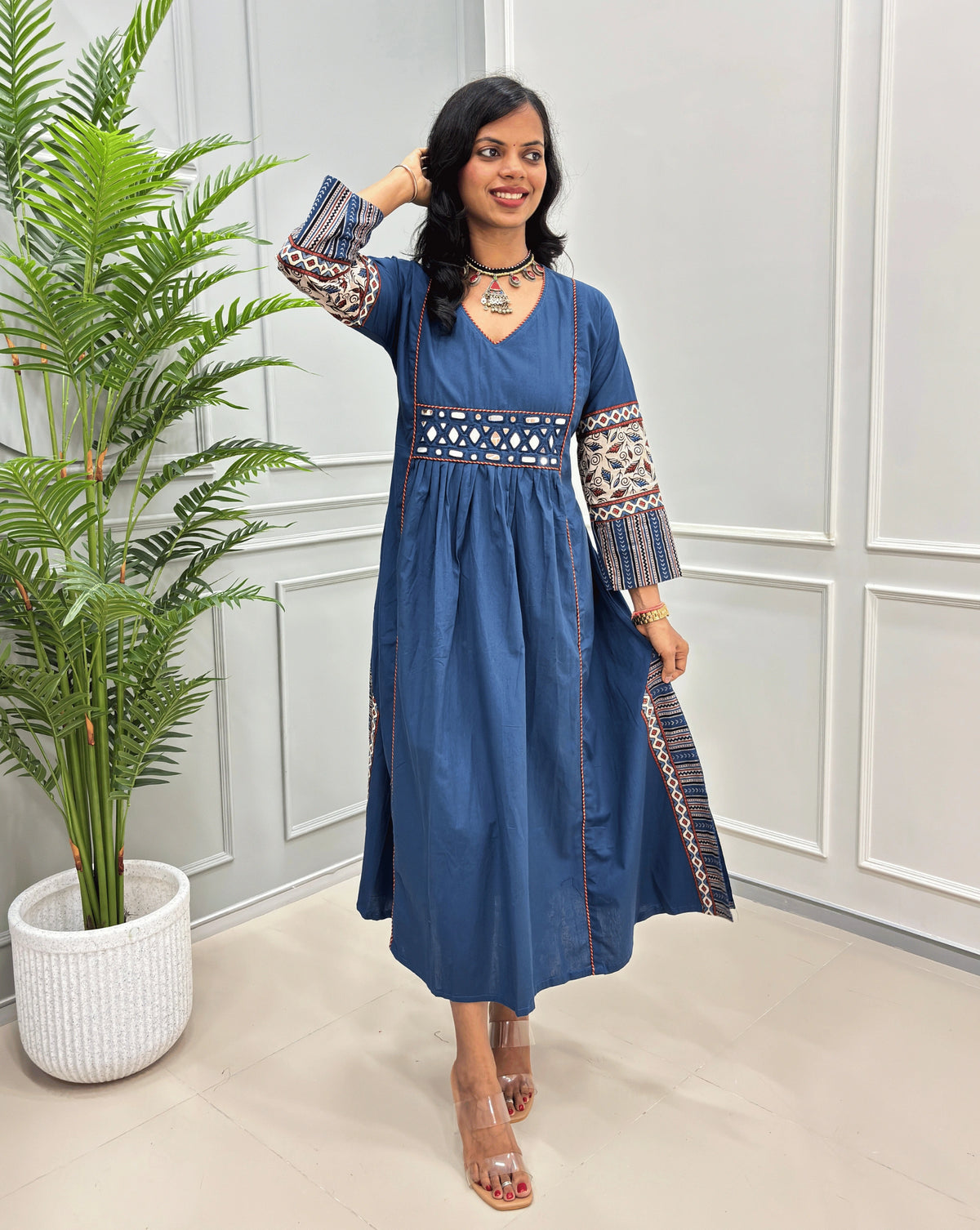 Blue cotton long kurti for women featuring handcrafted mirror work and Ajrakh print with intricate multicolor embroidered patterns on the sleeves and hem, standing in a bright room with a potted plant.