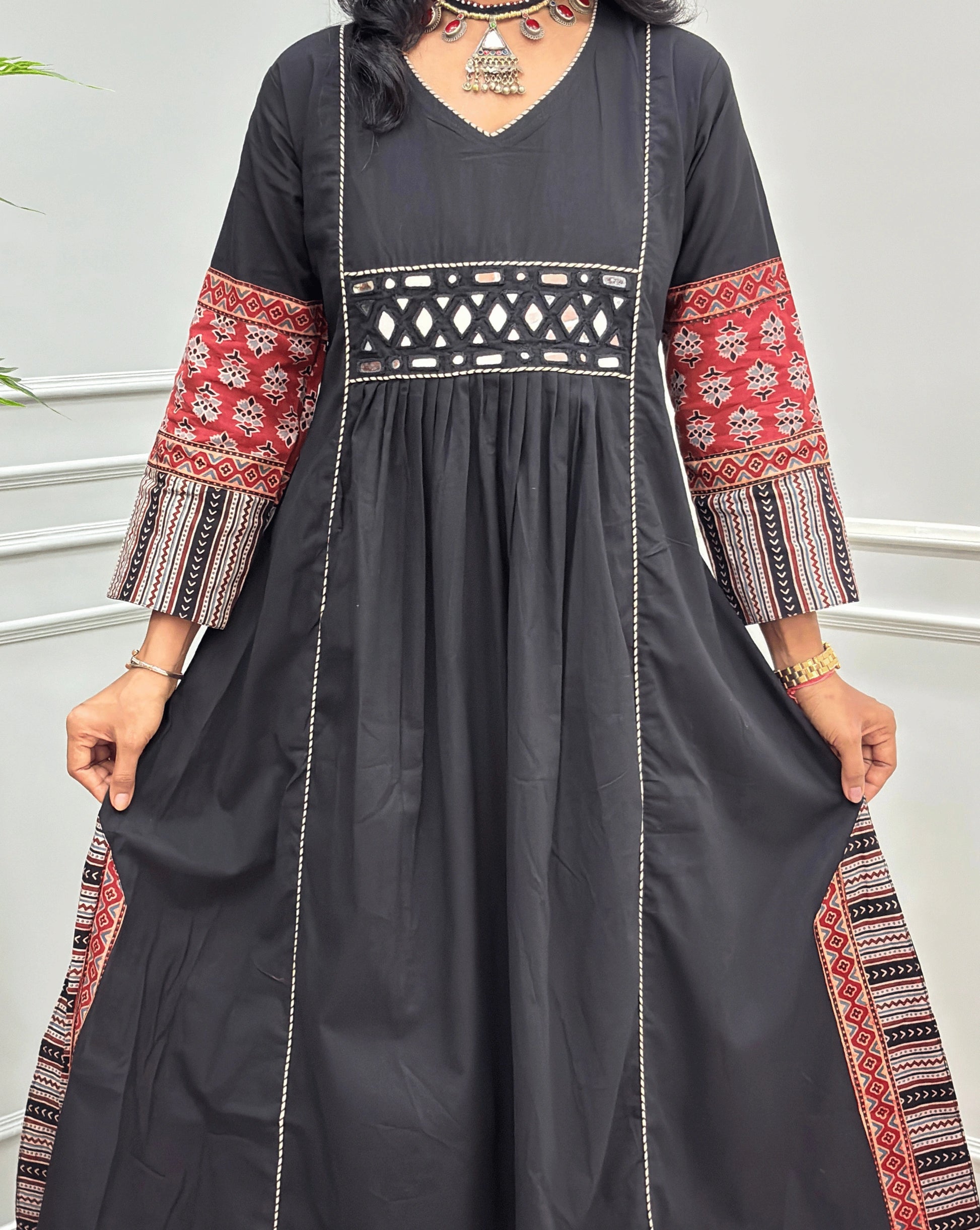 Close-up of handcrafted  black  Ajrakh cotton long kurti  with red and beige patterned sleeve cuffs, a matching patterned waistband, and white embroidered vertical lines, shown against a light grey indoor wall.