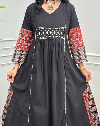 Close-up of handcrafted  black  Ajrakh cotton long kurti  with red and beige patterned sleeve cuffs, a matching patterned waistband, and white embroidered vertical lines, shown against a light grey indoor wall.