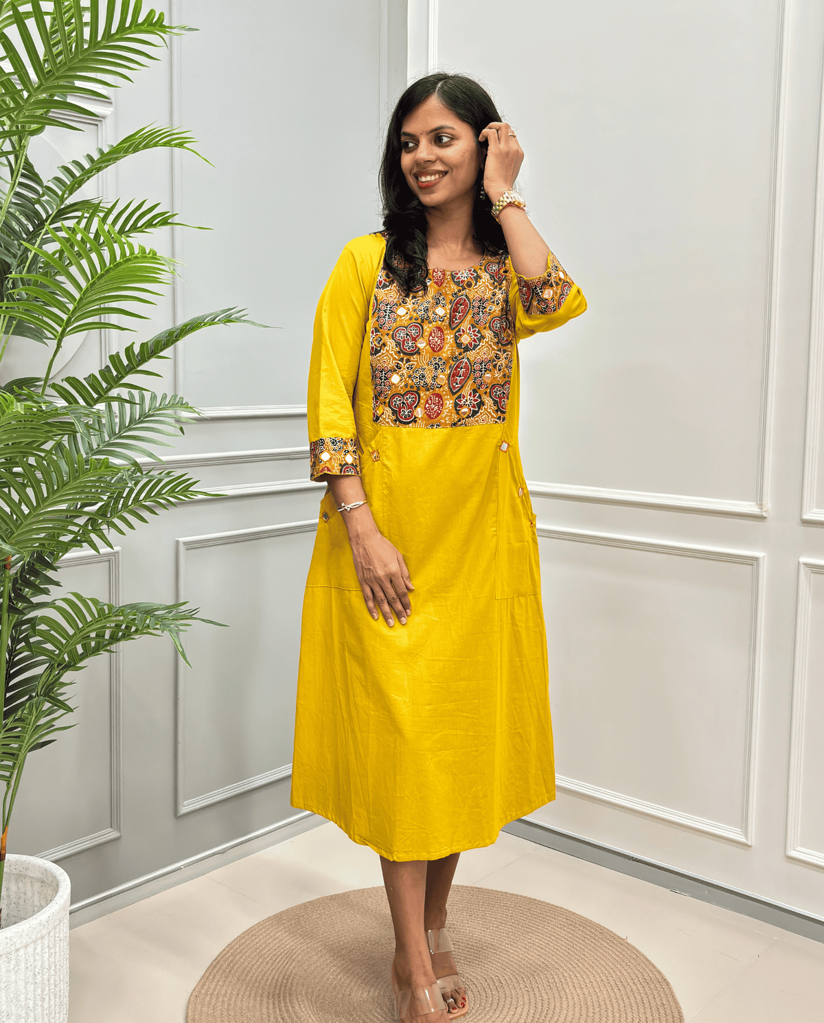 A woman wearing a Mustard Yellow A-line Cotton Long Kurti with a colorful embroidered yoke and patterned sleeve cuffs, standing in a room with white paneled walls and a potted plant.