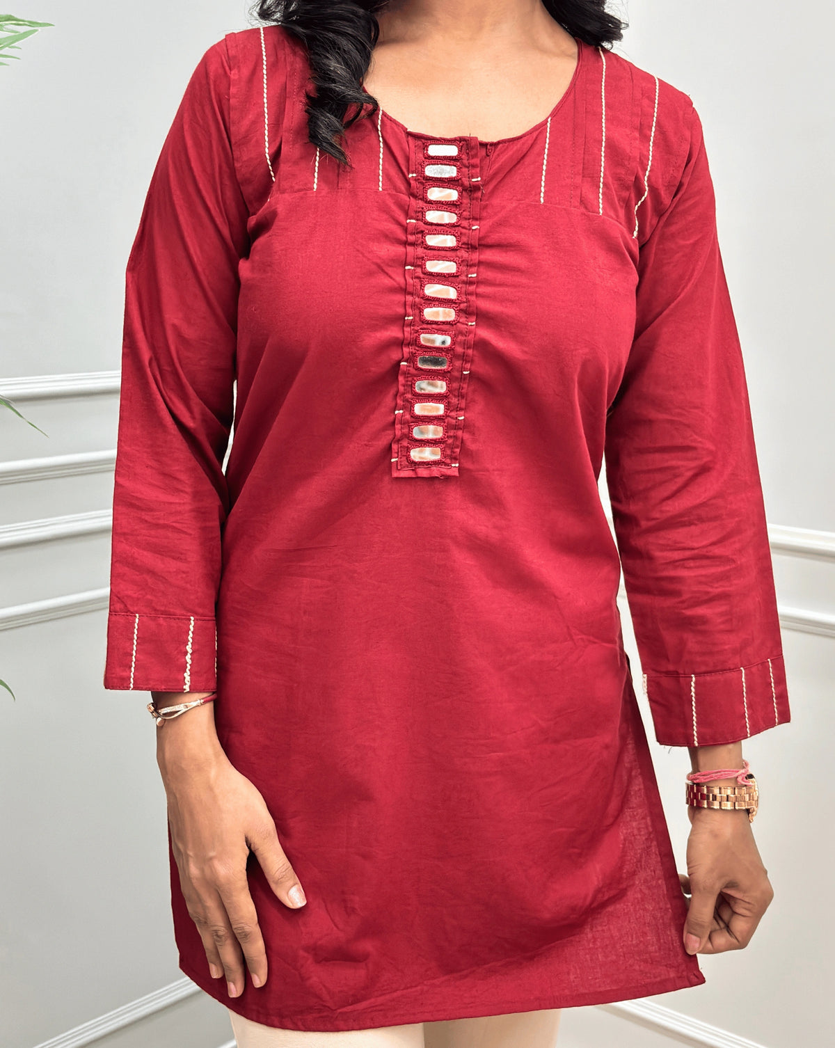 Close-up of Deep Red  women's Ajrakh Vegetable Dyed plant based Cotton Short-kurti with Beige piping and Hand-mirror work look a like button details, Round neck & Full sleeves, paired with beige Flared pants, standing in a room with a light gray paneled wall and a green plant on the left.