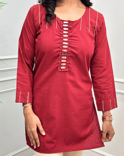 Close-up of Deep Red  women's Ajrakh Vegetable Dyed plant based Cotton Short-kurti with Beige piping and Hand-mirror work look a like button details, Round neck & Full sleeves, paired with beige Flared pants, standing in a room with a light gray paneled wall and a green plant on the left.