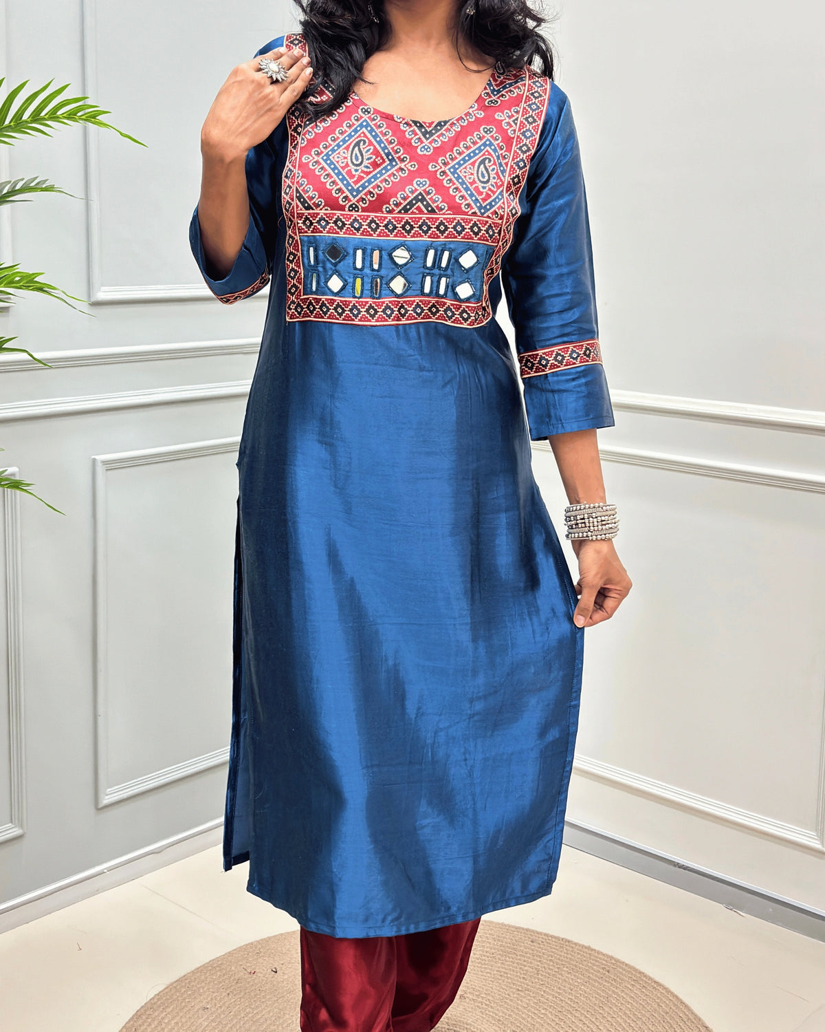 Close-up of Blue Ajrakh Mashru Silk Long-kurti with a highly detailed geometric embroidered yoke in blue, red, and white, along with ajrakh print border on front & sleeves, posed against a light gray paneled wall.