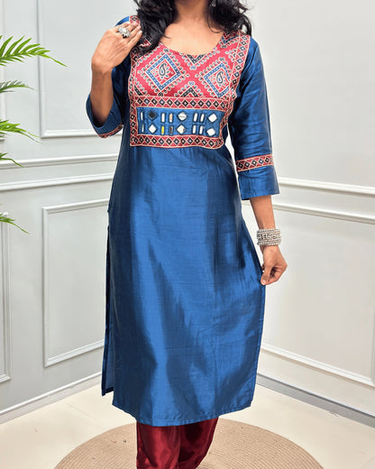 Close-up of Blue Ajrakh Mashru Silk Long-kurti with a highly detailed geometric embroidered yoke in blue, red, and white, along with ajrakh print border on front & sleeves, posed against a light gray paneled wall.