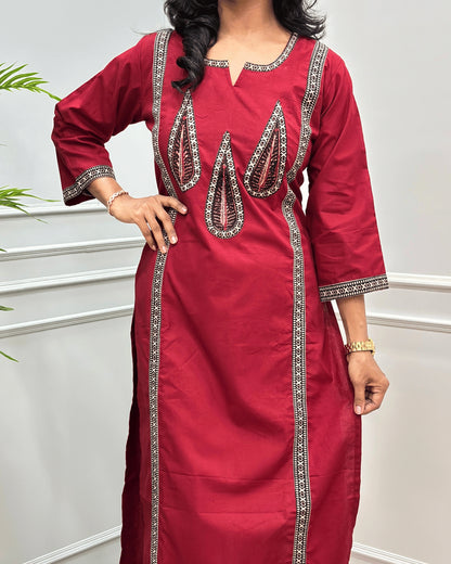 Close-up of Red Women's Hand-block  Ajrakh Pure Cotton Long-kurti with three-quarter sleeve featured with intricate Grey, Black and Red embroidered details on the neckline, cuffs, and front  panel, Along with Palm Leaf set against a light gray wall with a plant in the background.