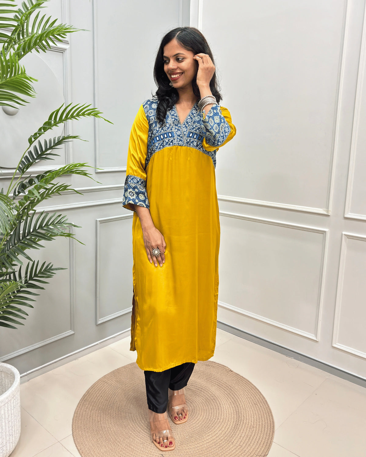 Mustard-Yellow Women's Ajrakh Modal Silk Long-Kurti with intricate Blue base & White Ajrakh print on the yoke and cuffs, paired with black pants and clear heeled sandals, standing on a round woven mat in a decorated room.