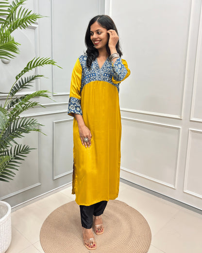 Mustard-Yellow Women's Ajrakh Modal Silk Long-Kurti with intricate Blue base & White Ajrakh print on the yoke and cuffs, paired with black pants and clear heeled sandals, standing on a round woven mat in a decorated room.