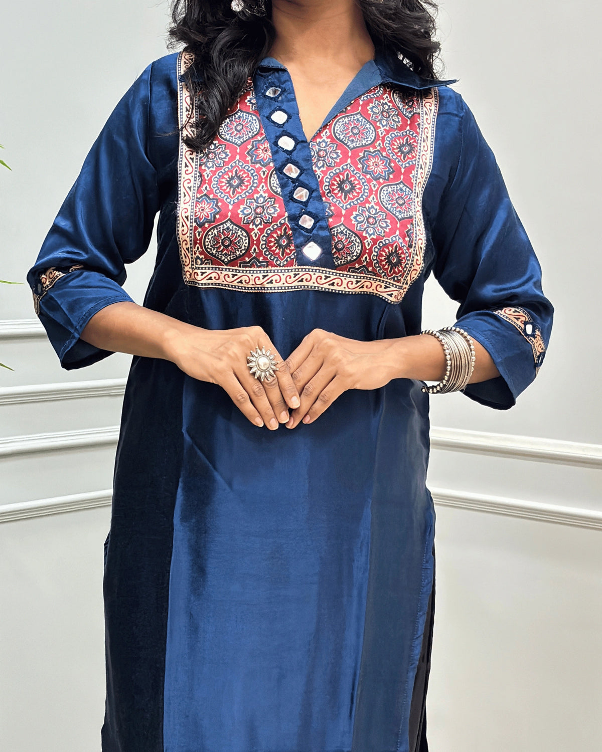 Close-up of Blue Ajrakh Mashru Silk Long-kurti with a heavily embroidered Badam-buta yoke and patterned sleeves, Hand-mirror Work on Front & Sleeves, Collar V- neck,  standing against a light gray paneled wall.