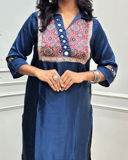 Close-up of Blue Ajrakh Mashru Silk Long-kurti with a heavily embroidered Badam-buta yoke and patterned sleeves, Hand-mirror Work on Front & Sleeves, Collar V- neck,  standing against a light gray paneled wall.