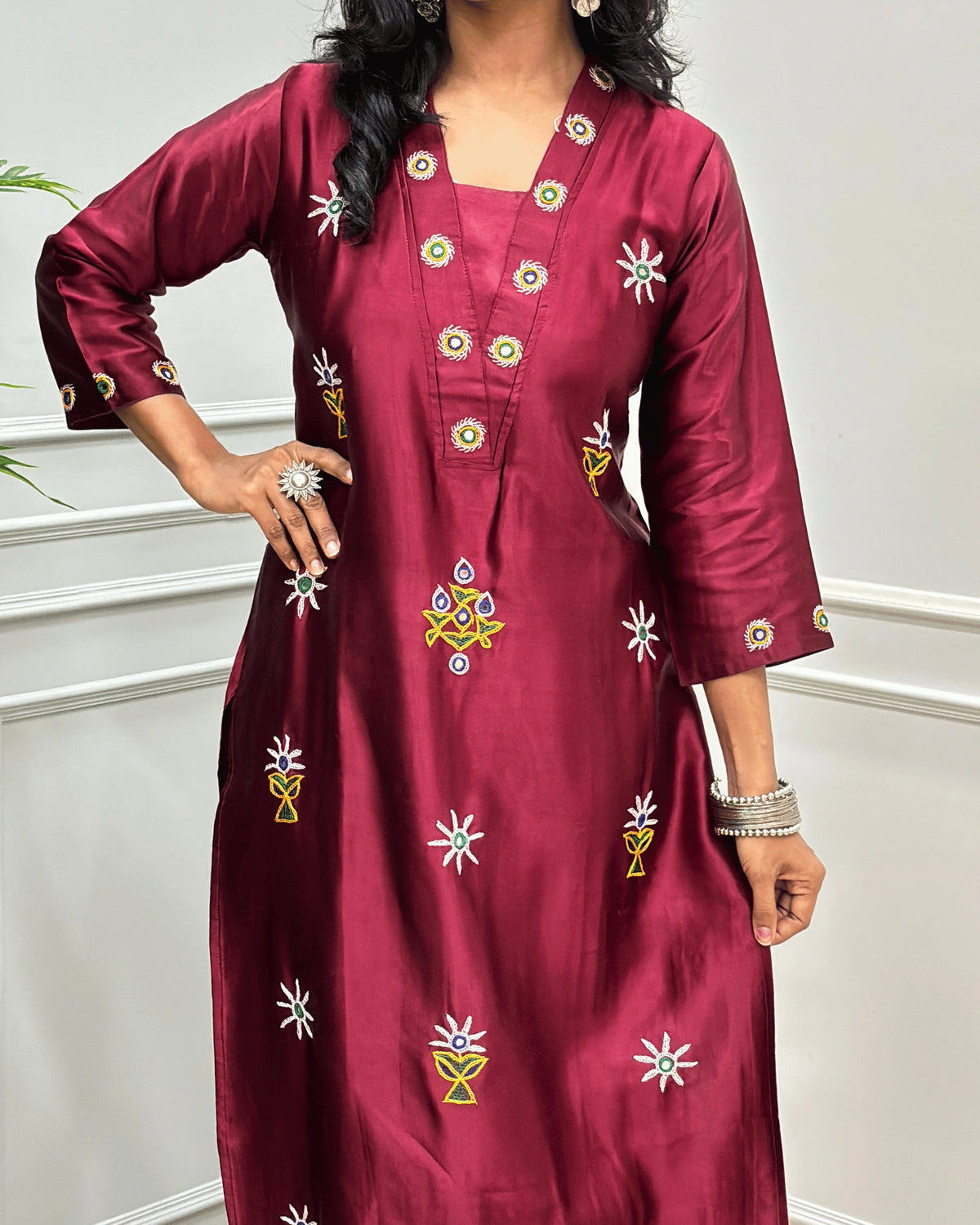Close-up view of Maroon Mashru Silk Long Kurti with colorful embroidered floral motifs and a Hand-mirror work on V-neckline, Front & Cuffs, Full Sleeves ,  standing against a light gray paneled wall. 