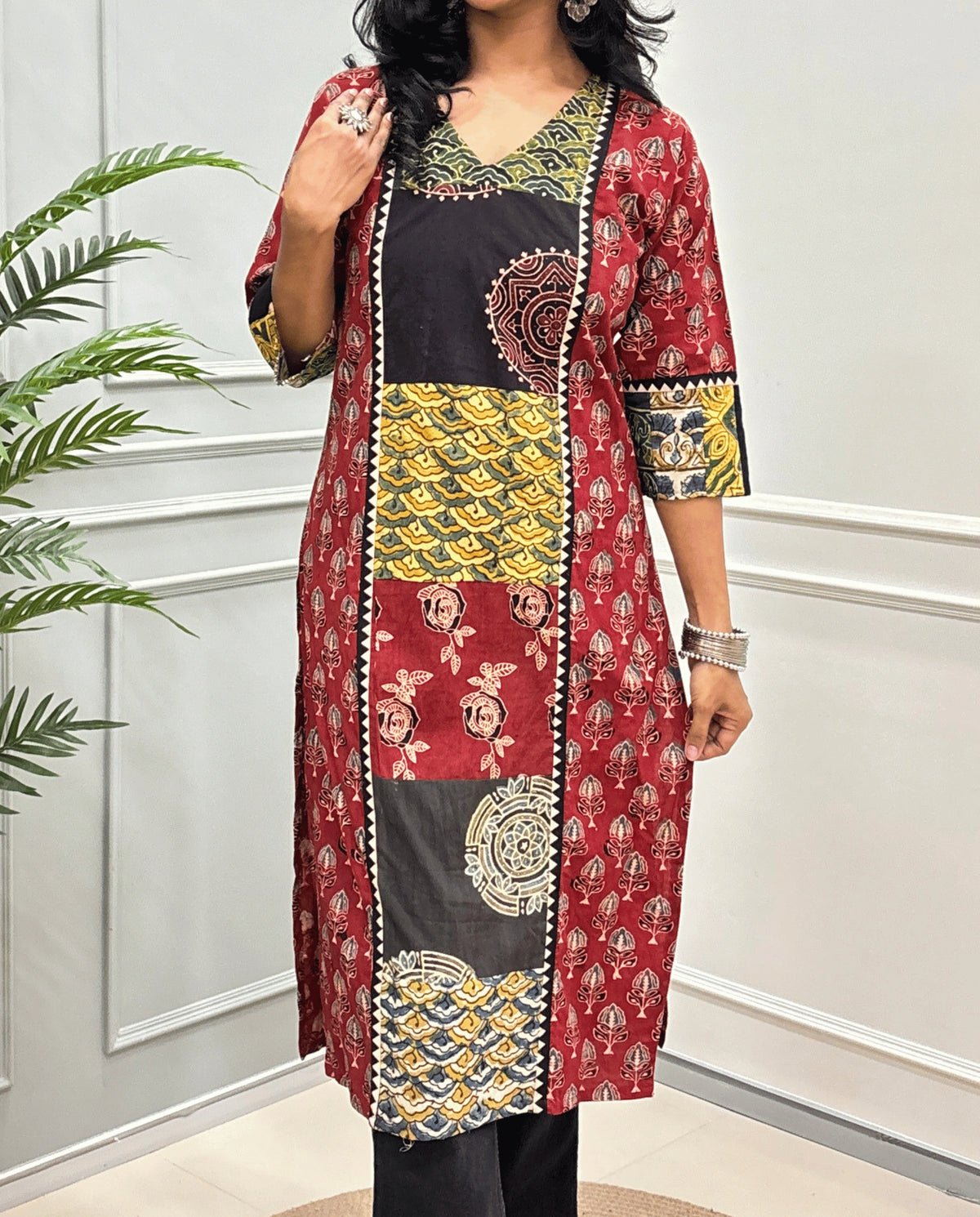 Close-up of Red Women pure cotton long kurti featured with blue, yellow, red, and black Geometric different print panels on Front & Sleeves cuffs , Ajrakh print on blue base outside the middle panel , three quarter Sleeves & V-neck standing indoors against a light paneled wall with a green plant on the left.