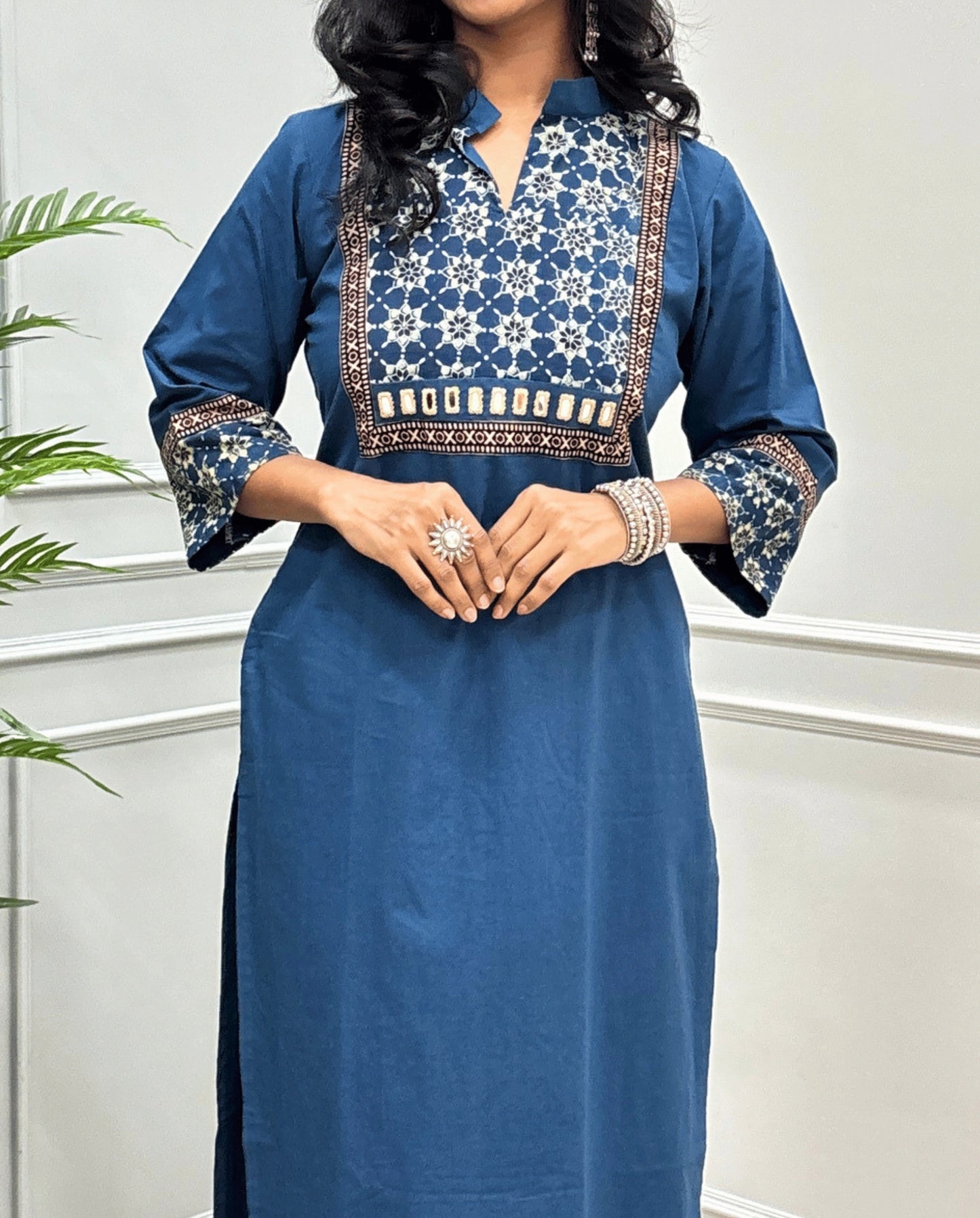 Close-up of Blue Hand-block Ajrakh Pure cotton Long kurti with three quarter sleeves with printed cuffs,  intricate black & beige geometric embroidery, with hand mirror work and thread work on border of mirror , with Mandarian collar V-neck and sleeve cuffs, standing against a light gray paneled wall.