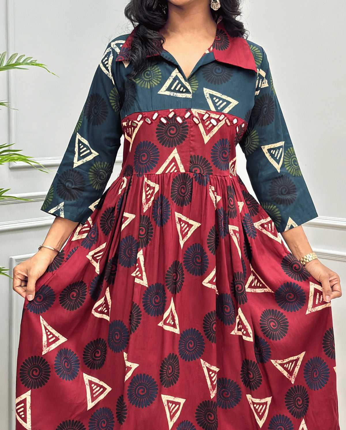 Deep Red Women's Ajrakh Vegetable dyed plant based Cotton Long-Kurti Featured with Three-Quarter Sleeves with a Green neck and cuffs, and a Deep Red-colored collar & dress featuring a geometric pattern of triangles and spiral in Beige, black, and blue, Hand-mirror work on front, standing on a round woven rug in front of a white paneled wall with a green potted plant on the left.