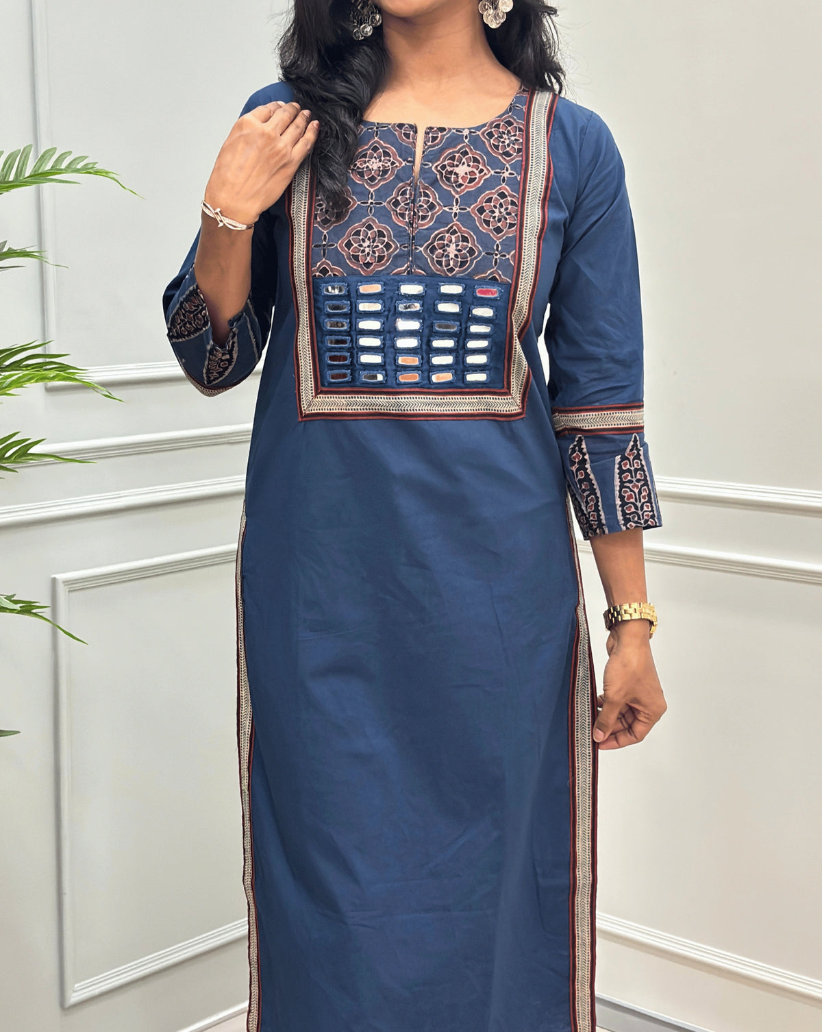 Close-up of Blue Women's Hand-block Ajrakh Pure Cotton Long Kurti Featured with Three-quarter Sleeves & Round neck with V-shape cut with a Blue base and abstract Black, Red & Beige geometric embroidery on the chest and sleeve cuffs, Hand-mirror work on Front, Ajrakh print border on Sides & Front, with side pockets, paired with black Flared pants, standing in a decorated room with a potted plant.
