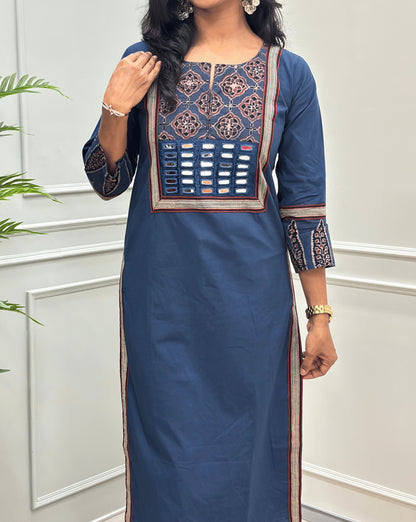 Close-up of Blue Women's Hand-block Ajrakh Pure Cotton Long Kurti Featured with Three-quarter Sleeves & Round neck with V-shape cut with a Blue base and abstract Black, Red & Beige geometric embroidery on the chest and sleeve cuffs, Hand-mirror work on Front, Ajrakh print border on Sides & Front, with side pockets, paired with black Flared pants, standing in a decorated room with a potted plant.