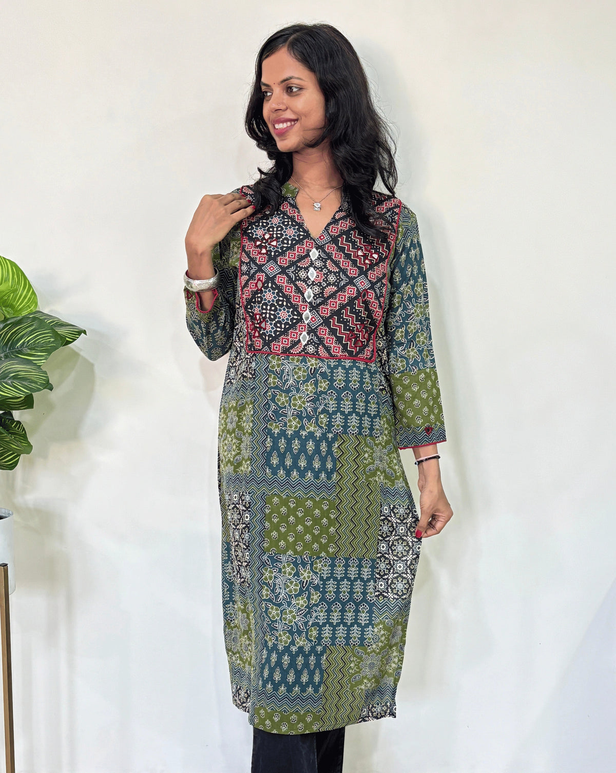 Front view of Green Women's Ajrakh Vegetable Dyed plant based cotton long kurti with green, black, and red patterns, Hand-mirror work on front & Sleeves paired with black flared pants, standing in a room with a white wall and a potted plant.