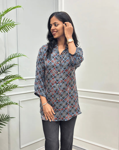 Front View of Blue Women's Ajrakh Pure Modal Silk Shirt with a Floral print on blue, Black, and Red geometric pattern, black flared pants, standing on a round woven rug in front of a white paneled wall with a potted plant on the left