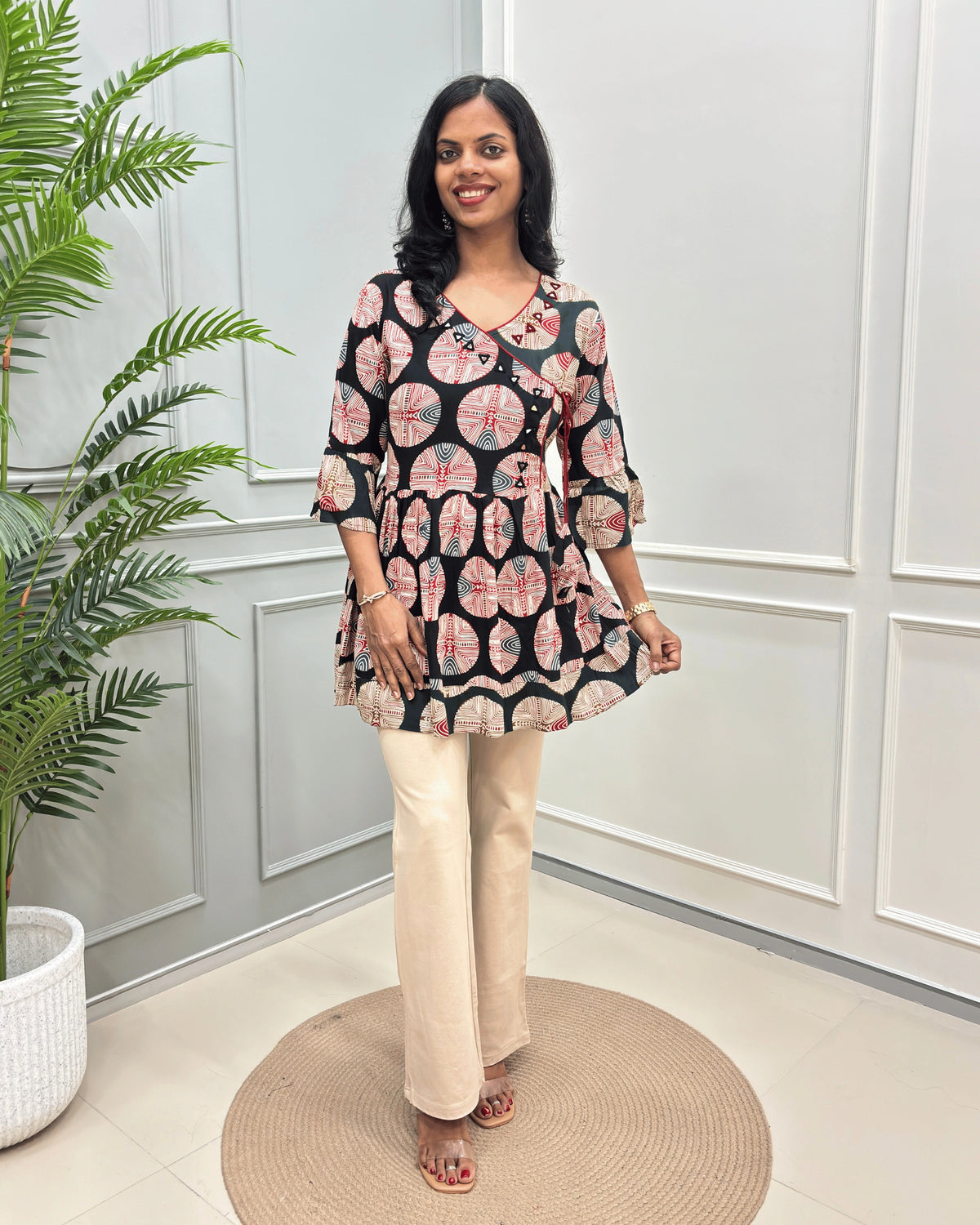 Black Women's Ajrakh Vegetable dyed plant based Cotton Short-Kurti with three-quarter frill sleeves, and waist to bottom,  paired with beige flared pant , standing on a round woven rug in front of a white paneled wall with a potted plant on the left.