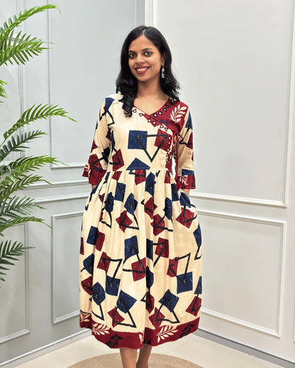 Front View of Beige women's plant based rayon cotton long-kurti with a geometric pattern in navy-blue,  deep red & black, 2 side pockets with frill sleeves , standing in front of a white paneled wall with a green plant on the left.