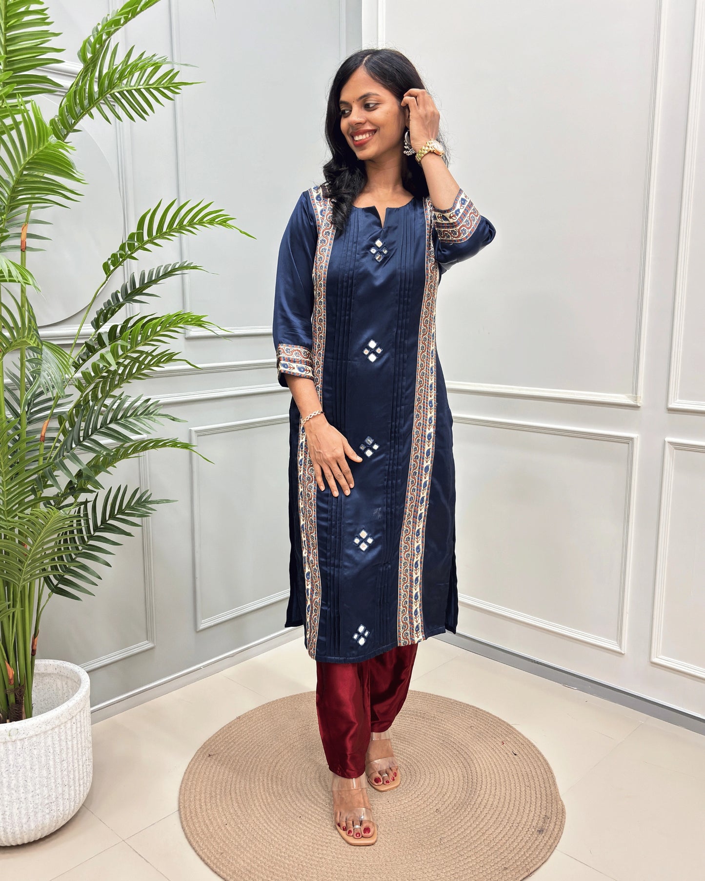Navy-Blue women's Ajrakh Mashru Silk long kurti with beige and white geometric border details, paired with maroon pants, standing on a round woven mat in front of a white paneled wall with a potted plant.