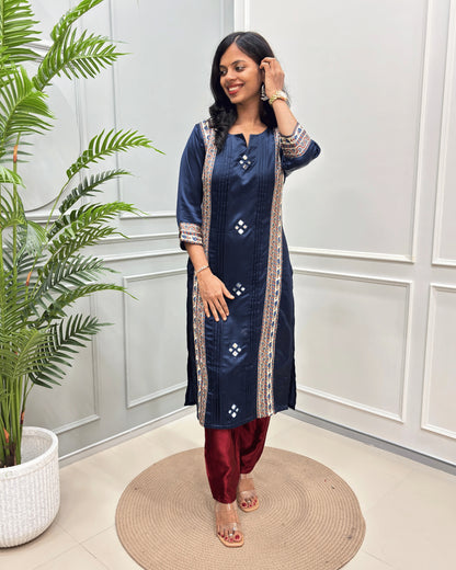 Navy-Blue women's Ajrakh Mashru Silk long kurti with beige and white geometric border details, paired with maroon pants, standing on a round woven mat in front of a white paneled wall with a potted plant.