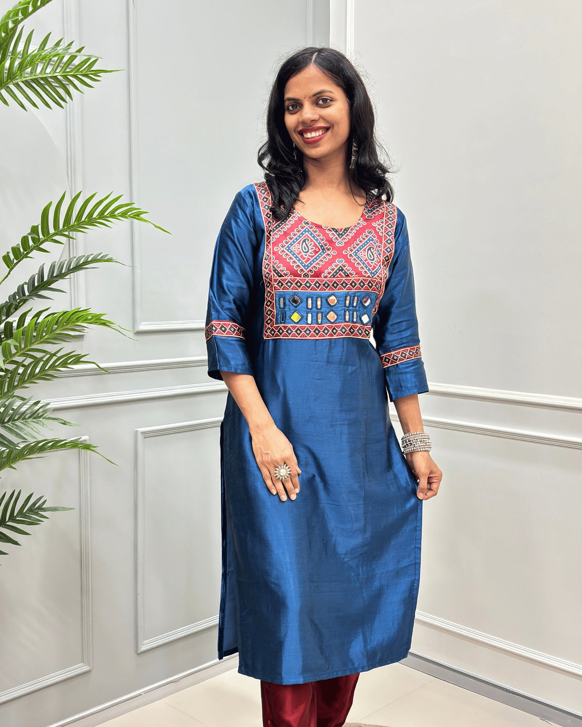 Front view of Blue Ajrakh Mashru Silk Long-kurti with a blue, red, and white in Bandhani print , featuring Hand-mirror work with Yellow thread work embellishments , blue & red patterned cuffs, standing against a white paneled wall.