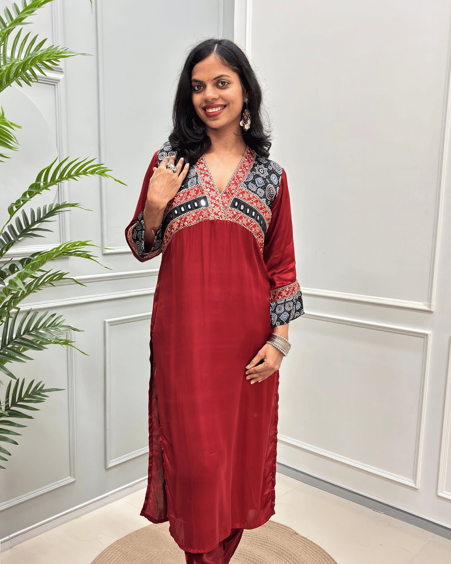 Front view of Deep-Red women's Ajrakh Modal Silk Long kurti featured with Black base & ajrakh print on neck & cuffs , hand-mirror work on chest standing in a white paneled room.