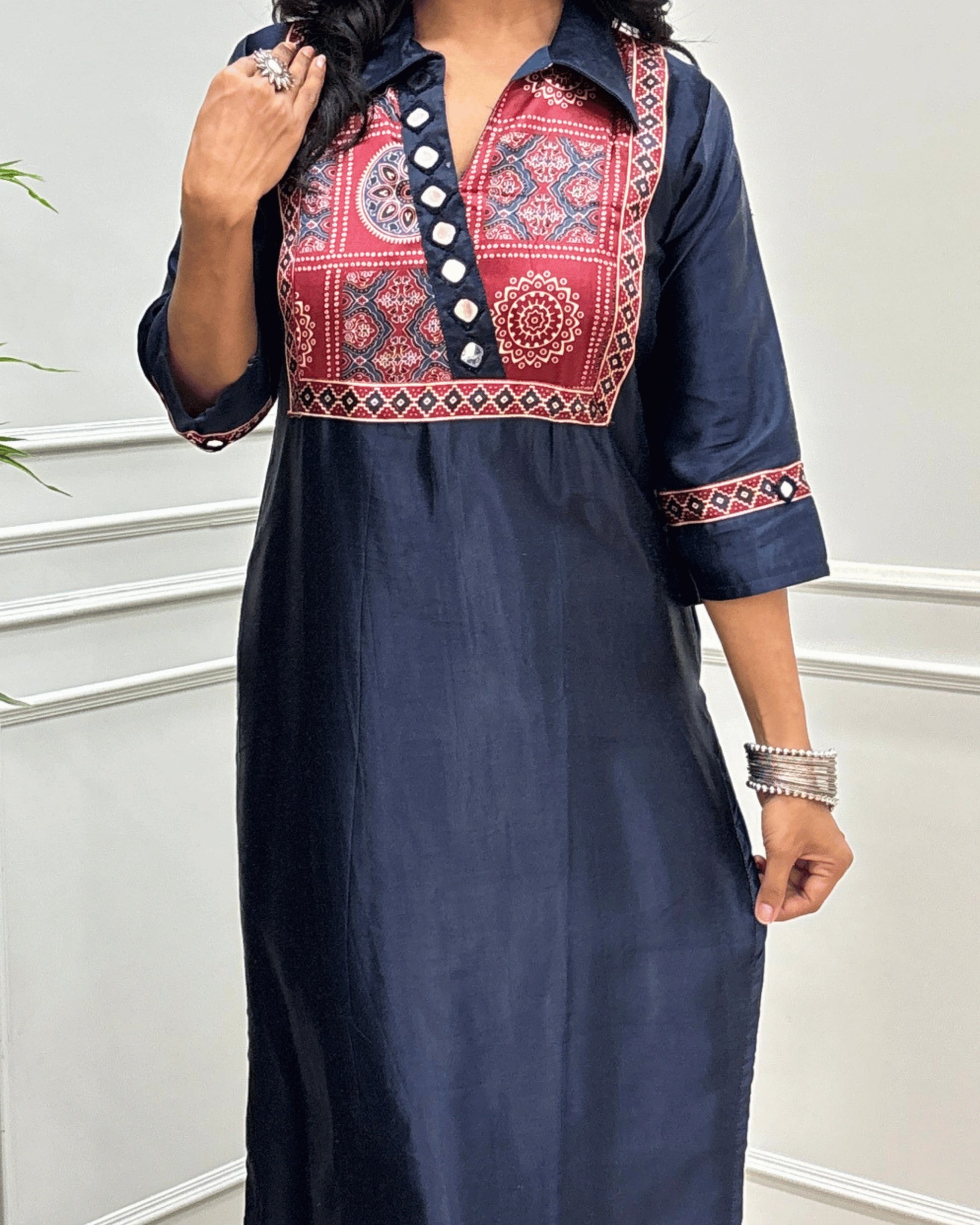 Close-up of Navy-Blue Ajrakh Mashru Silk Long-kurti with a heavily embroidered Ajrakh yoke and patterned sleeves, Hand-mirror Work on Front & Sleeves, Collar V- neck,  standing against a light gray paneled wall.