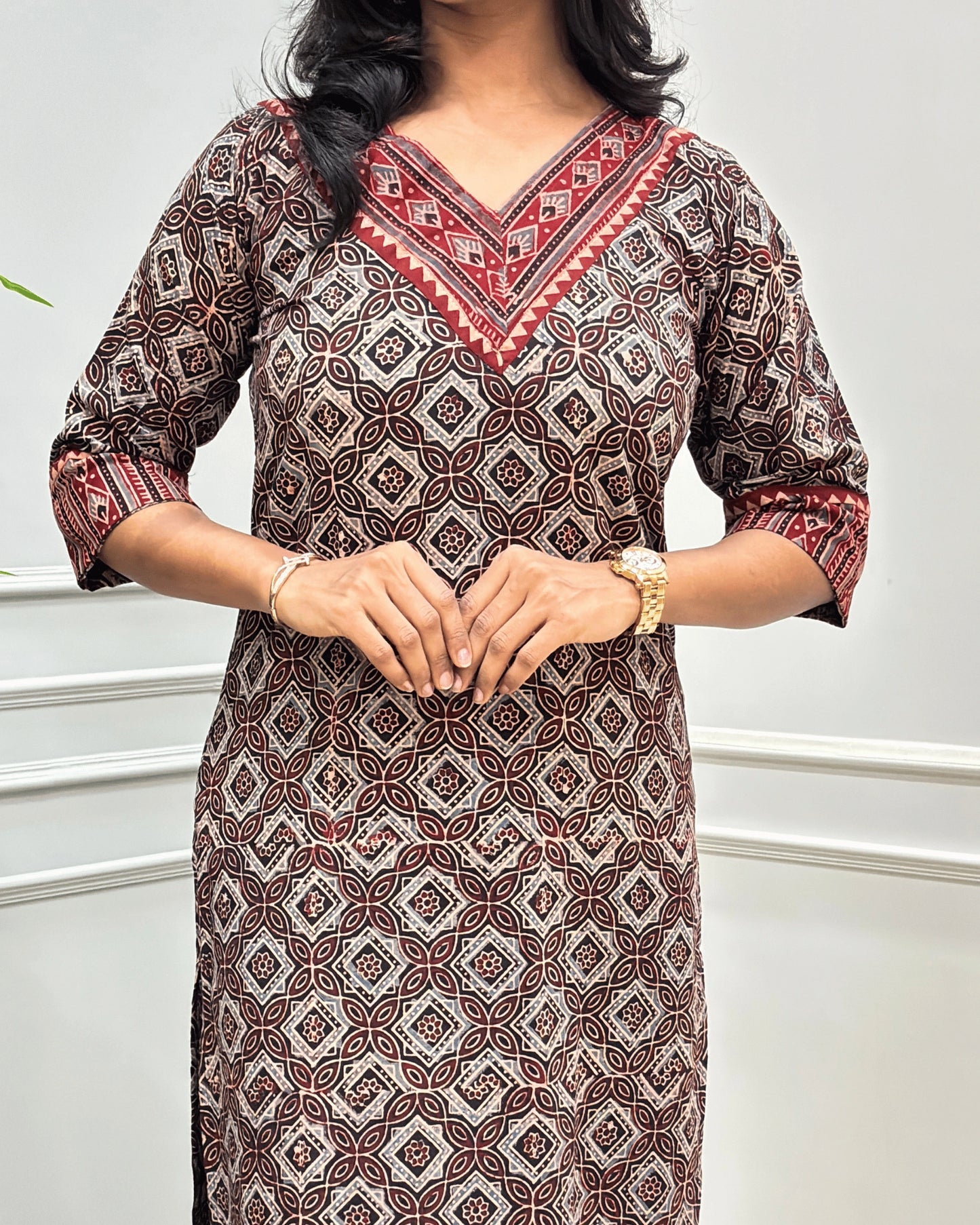 Close-up of brown and red Hand-block Ajrakh pure cotton Long-kurta with a Ajrakh geometric pattern, featuring a V-neckline and patterned sleeve cuffs, standing against a white paneled wall.
