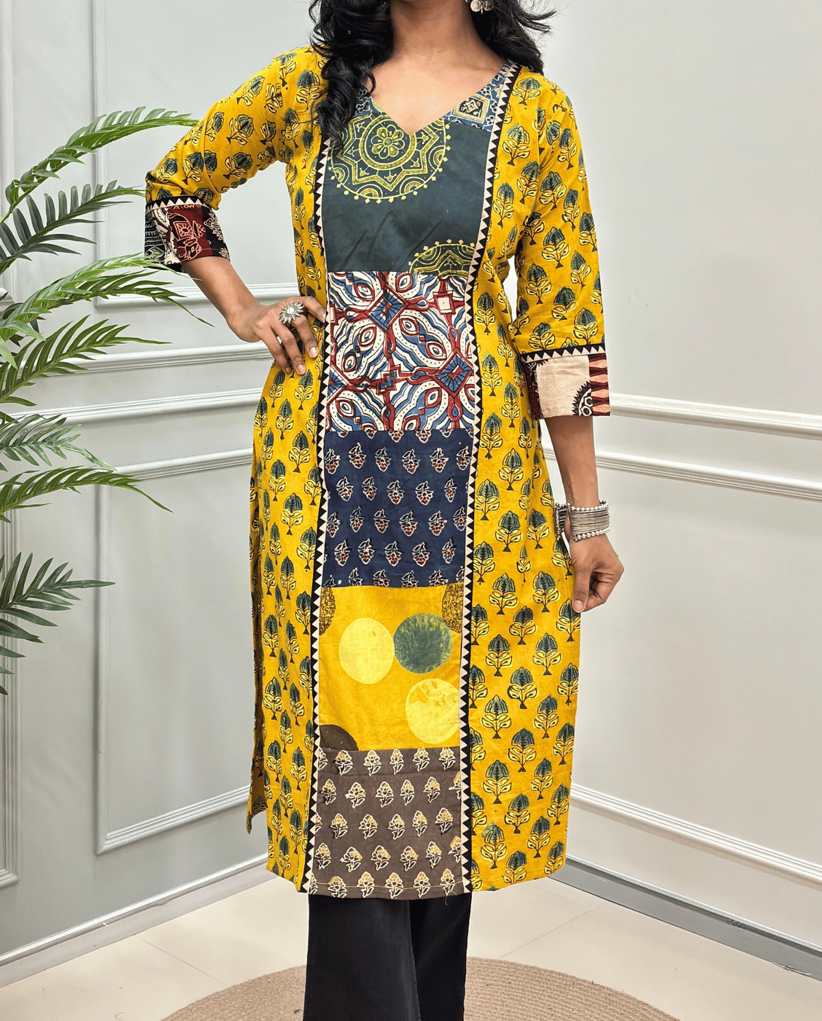 Close-up of Yellow Women pure cotton long kurti featured with blue, yellow, red, and black Geometric different print panels on Front & Sleeves cuffs , Ajrakh print on blue base outside the middle panel , three quarter Sleeves & V-neck standing indoors against a light paneled wall with a green plant on the left.