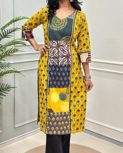 Close-up of Yellow Women pure cotton long kurti featured with blue, yellow, red, and black Geometric different print panels on Front & Sleeves cuffs , Ajrakh print on blue base outside the middle panel , three quarter Sleeves & V-neck standing indoors against a light paneled wall with a green plant on the left.